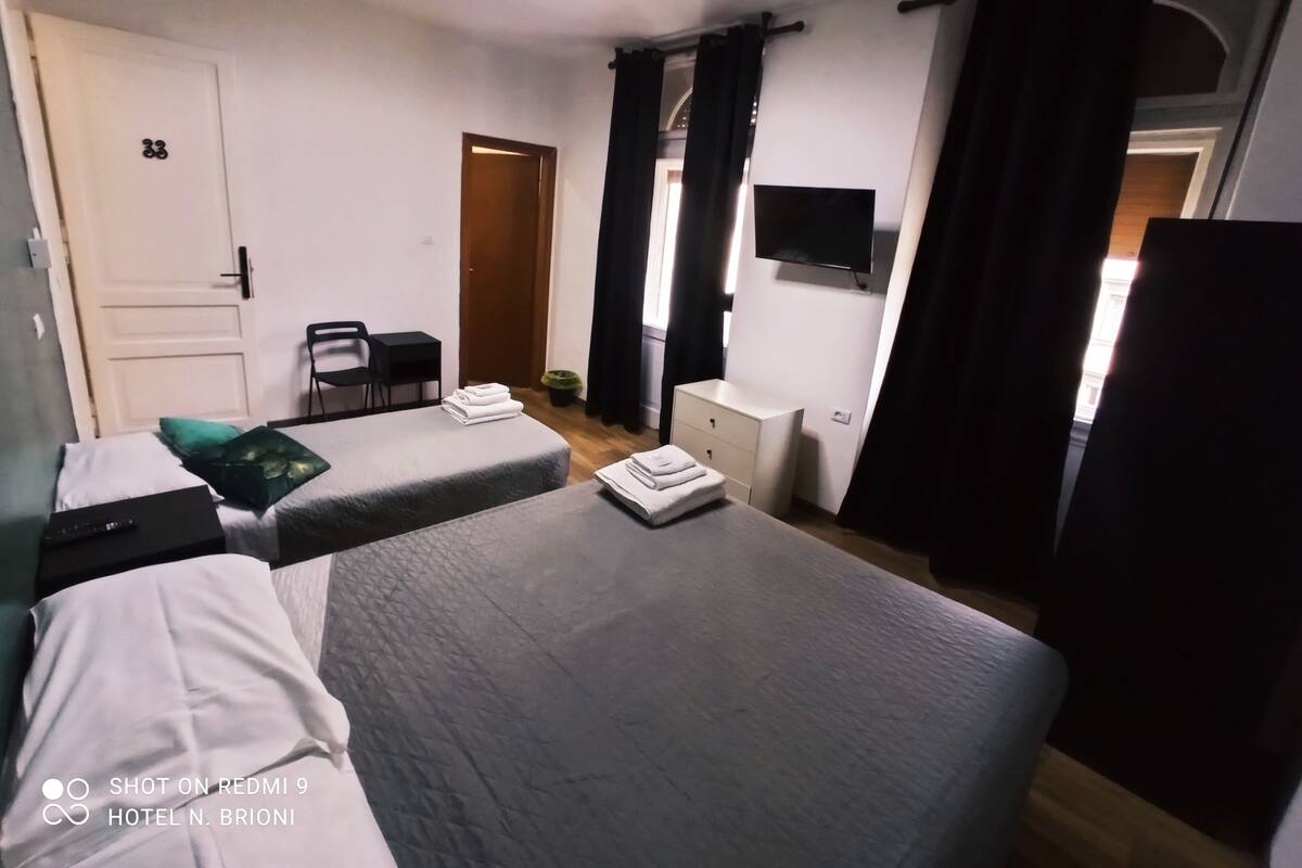 A hotel room with two beds, a desk, a chair, a TV, and a door with a number.