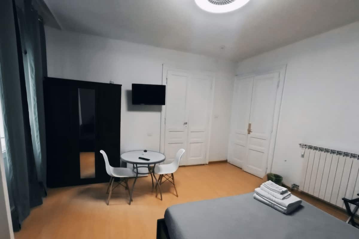 A small, clean room with a bed, a desk, two chairs, a TV, and a radiator.