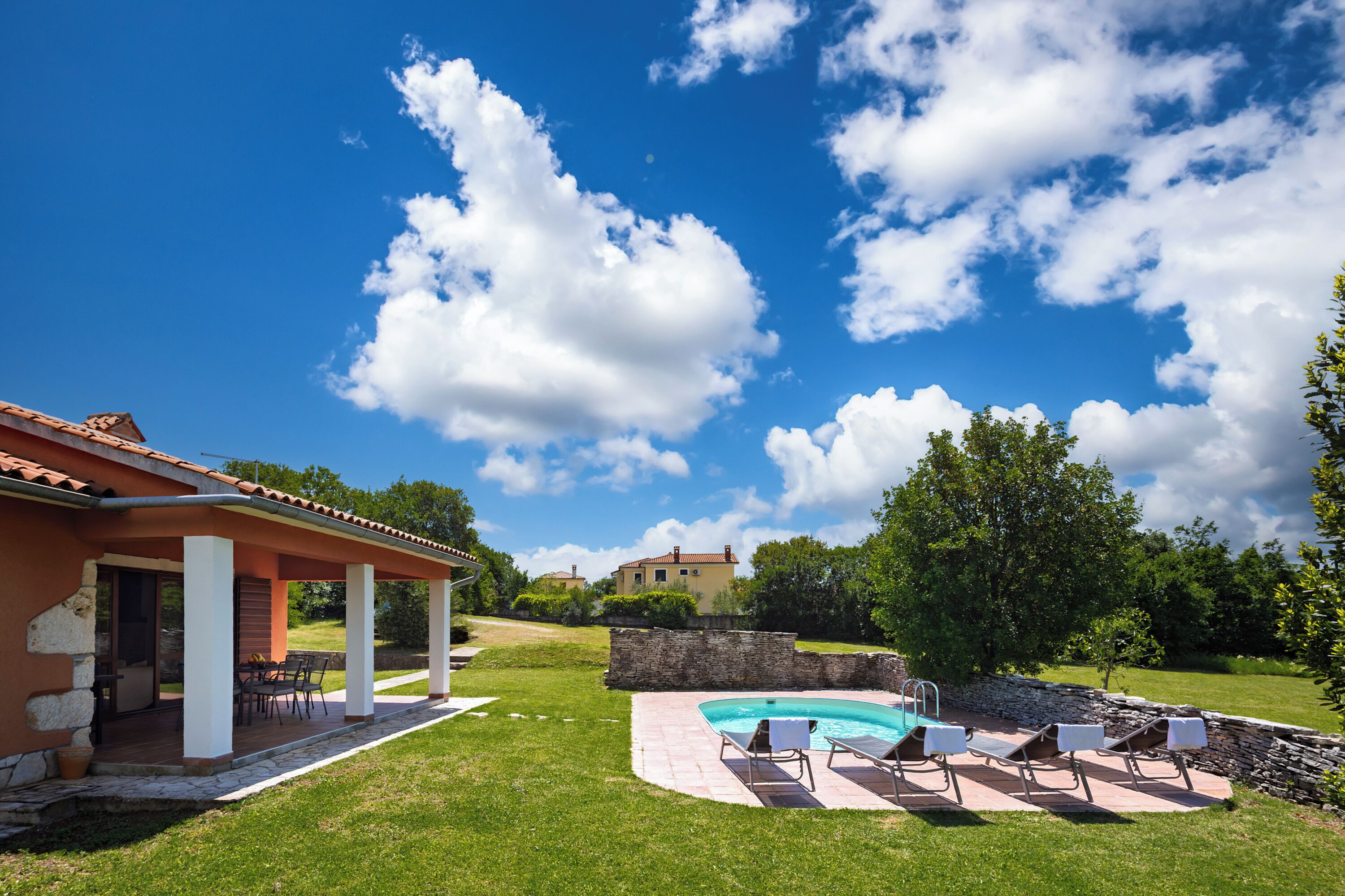 Una zona junto a la piscina con sillones de descanso, un porche cubierto y un muro de piedra.