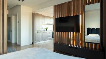 Una habitación de hotel moderna con una cama grande, un televisor de pantalla plana montado en un panel de madera, y vistas a la cocina y al pasillo.