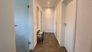 Um corredor estreito com chão de azulejos, paredes brancas e uma pequena mesa com cadeiras.