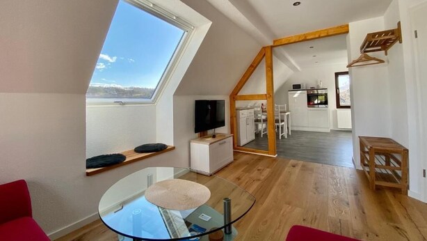 Una sala de estar moderna con una mesa de vidrio, un sillón rojo, un televisor y vista a la cocina a través de una puerta abierta.