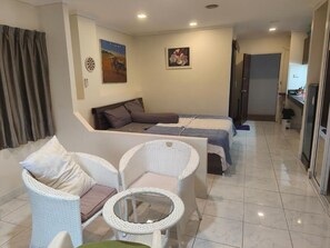 A room with a bed, two white wicker chairs, a small round table, and a sofa. There are two paintings on the wall, a door, and a kitchen area visible through an open door. - Jomtien Hill Resort Pratamnak 108 (Pattaya)