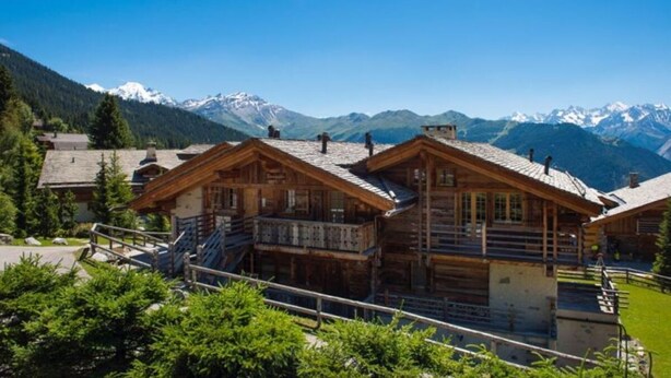 A wooden chalet with balconies, set against a backdrop of snow-capped mountains.