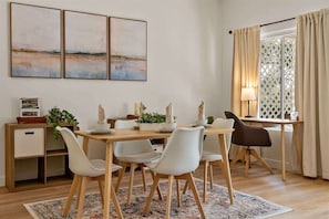 A dining area with a wooden table, white chairs, and a patterned rug. There are three framed artworks on the wall, a small plant on the side table, and a window with curtains.