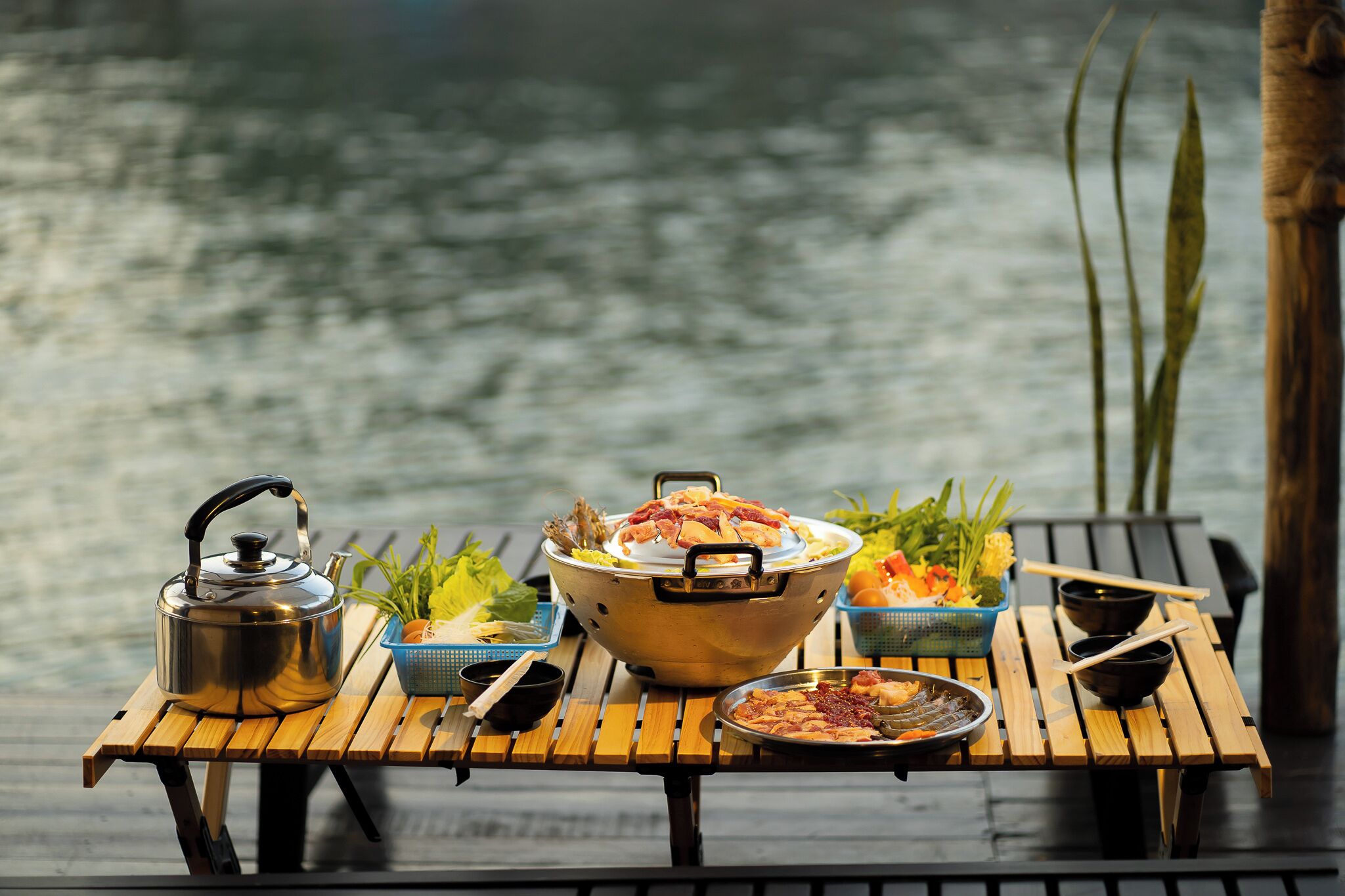 Una mesa de madera con una tetera, un bol grande y diferentes platos, con vistas al agua.