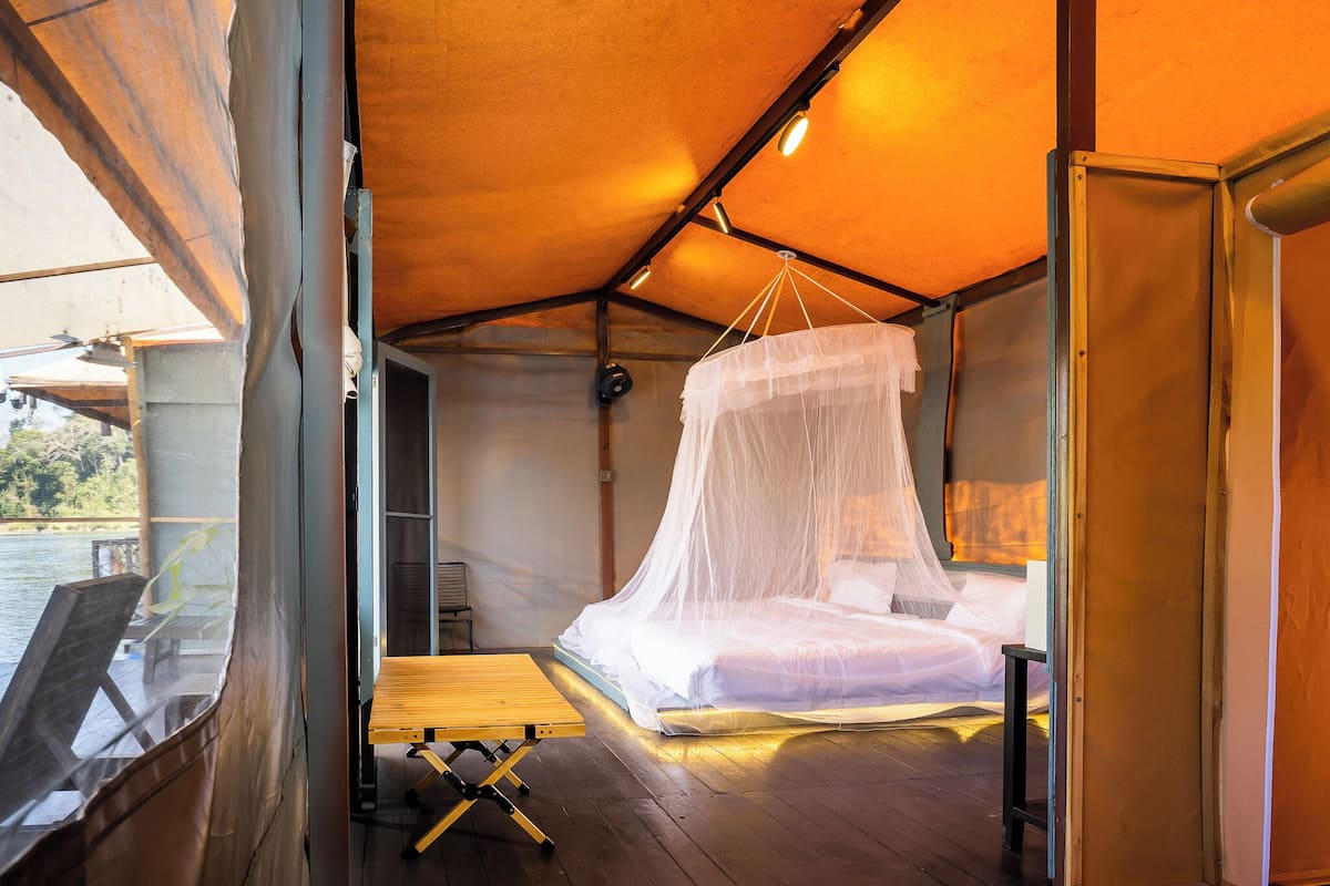 A tented room with a bed, a table, and a view of the water outside.