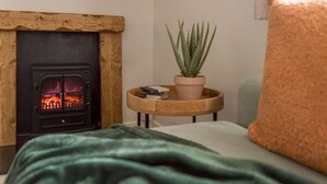 A cozy room with a wood-burning stove, a green blanket, a small round table with a plant, and a cushion. - Squirrel Bank, with a hot tub & parking (Bowness-on-Windermere)
