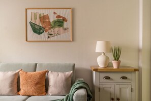 A room with a grey sofa, a framed abstract painting, a white lamp, and a potted plant on a wooden side table. - Squirrel Bank, with a hot tub & parking (Bowness-on-Windermere)