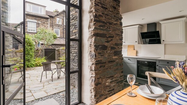 Une cuisine moderne aux murs en pierre, un coin repas avec une table et des chaises, et une vue sur un bâtiment en pierre à l’extérieur.