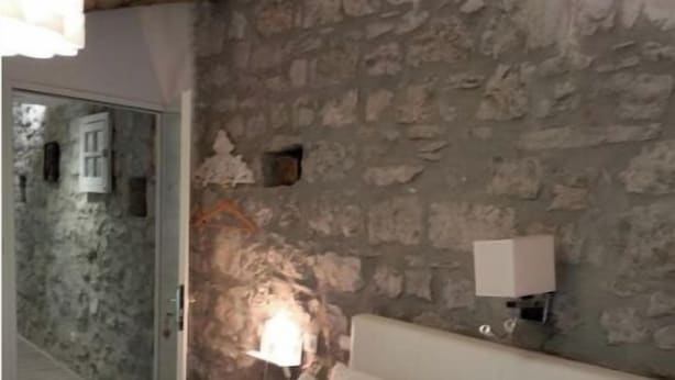 Un dormitorio con pared de piedra, una cama con ropa de cama blanca, una mesita de noche con una lámpara y una puerta que da a otra habitación.