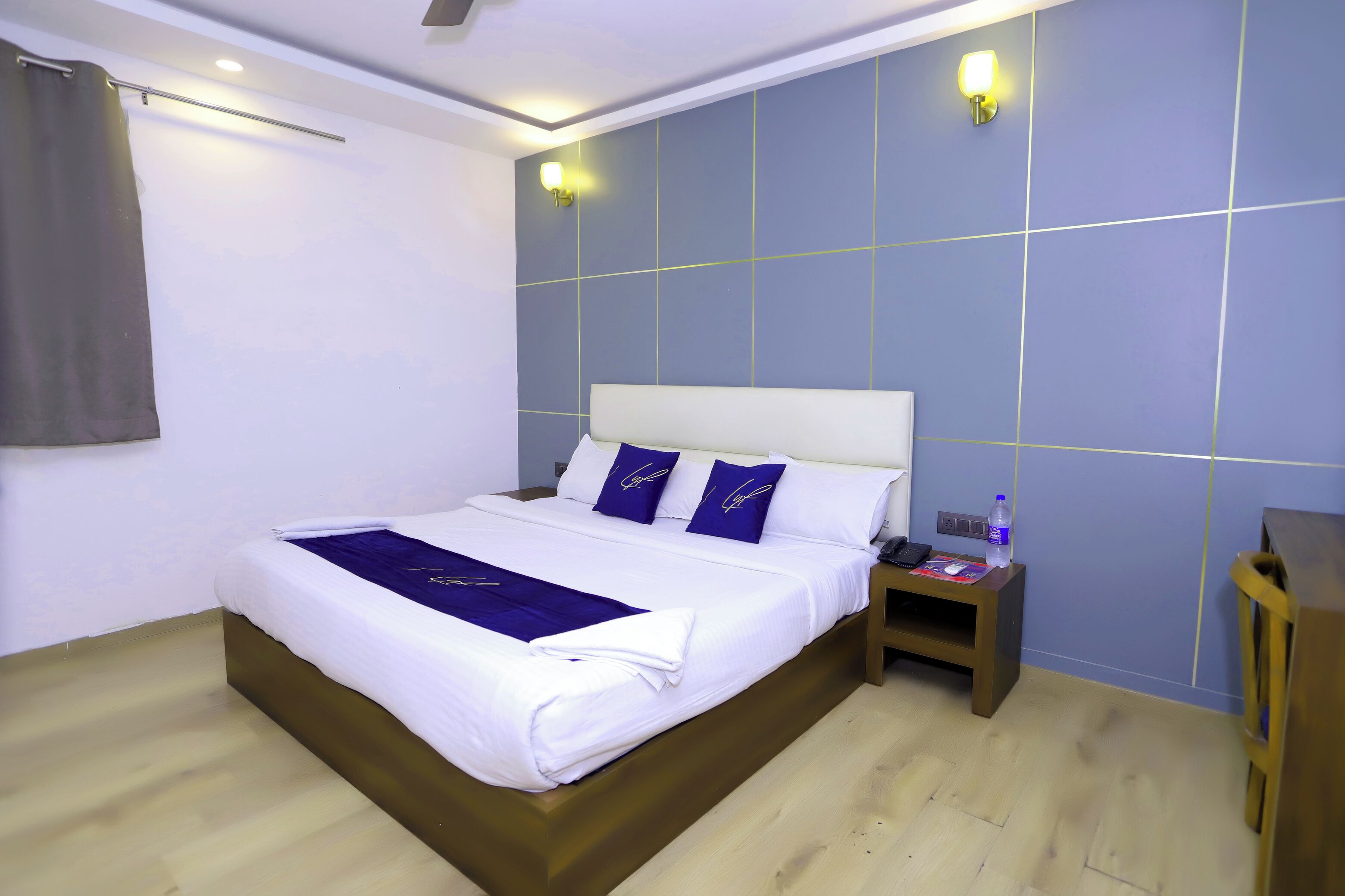 Un lit bien fait, avec du linge de lit blanc et violet, une table de chevet en bois avec une bouteille d’eau, et un luminaire fixé au mur.