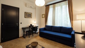 Una sala de estar con un sofá azul, un escritorio con una silla, una lámpara y un ventanal con cortinas.