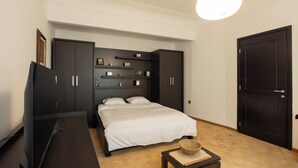Un dormitorio con cama, armario de madera oscura, estanterías con objetos decorativos, televisor y una puerta.