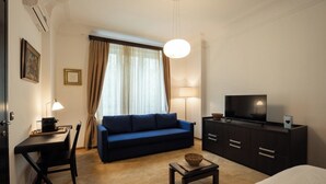 Habitación de hotel con un sofá azul oscuro, una mesita con una cesta, un televisor en un mueble y una cama.