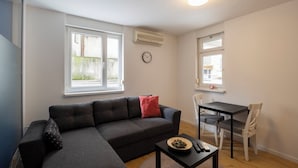 Un salon avec un canapé gris, une petite table sur laquelle se trouve un saladier rempli de fruits, une horloge et une fenêtre avec un climatiseur.
