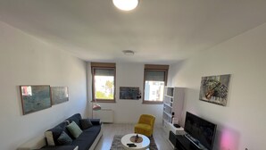 Un salon moderne avec un canapé, un fauteuil jaune, une table basse et un téléviseur. Il y a deux fenêtres équipées de stores, des œuvres d’art accrochées aux murs et un luminaire au plafond.
