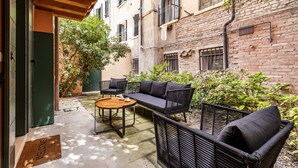 An outdoor seating area with wicker furniture and a wooden table, surrounded by greenery and a brick building. - The Blue Light by Wonderful Italy (Venecia)