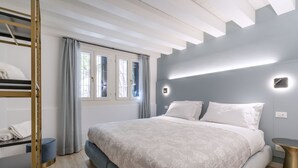 A modern bedroom with a bunk bed, a large bed with a patterned bedspread, a bedside table, and a window with curtains. - The Blue Light by Wonderful Italy (Venecia)