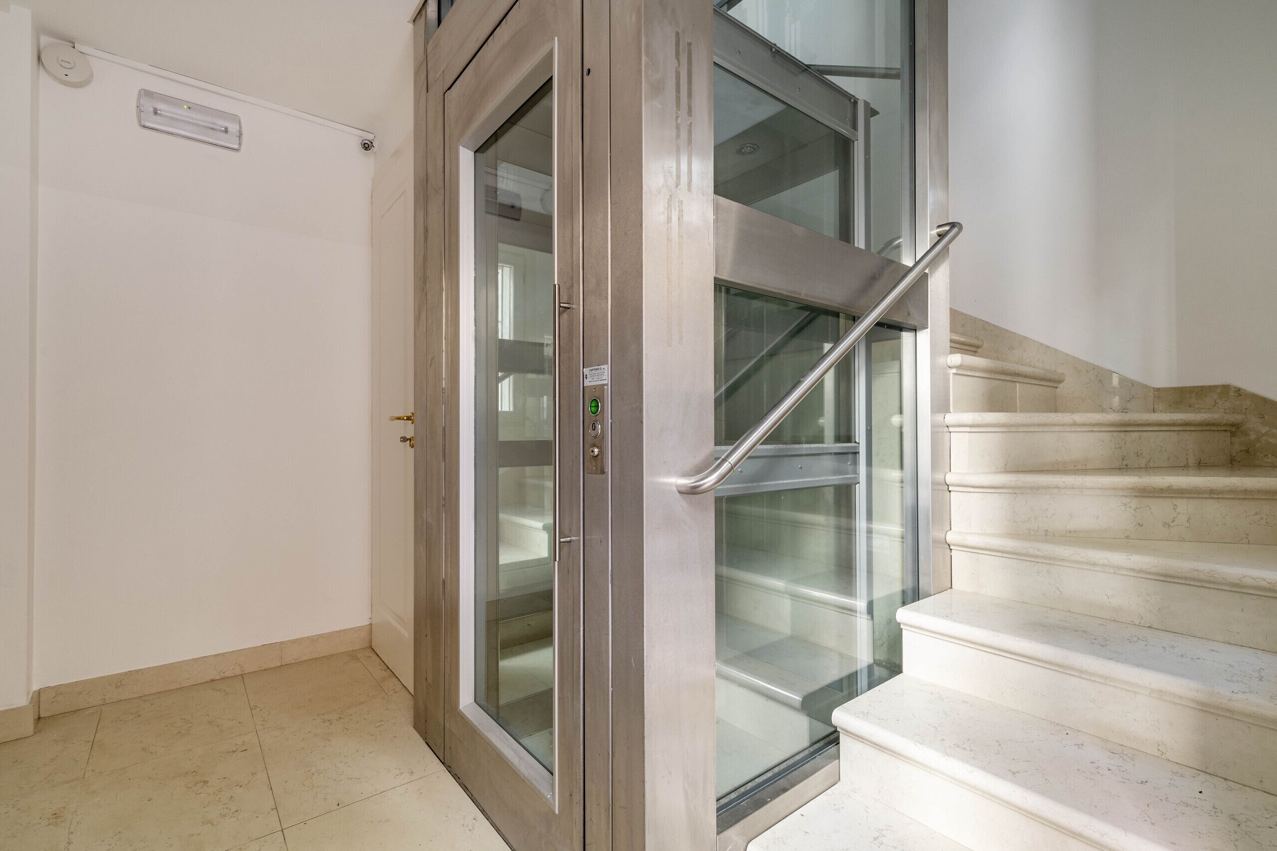 A modern elevator with a glass door and a metal handrail, adjacent to a marble staircase.