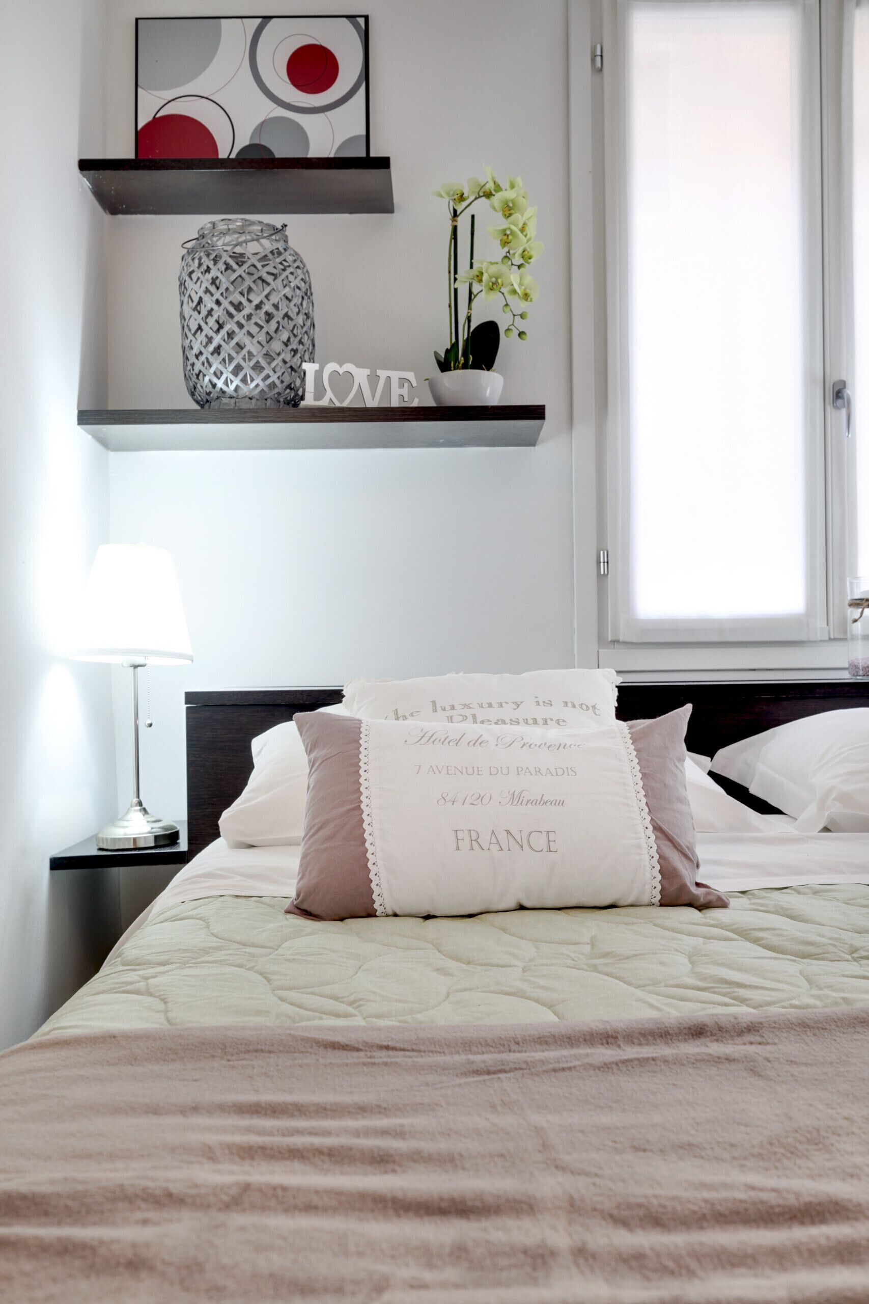 A neatly arranged hotel room with a bed, a bedside lamp, a decorative shelf with a vase and flowers, and a framed artwork above the shelf.