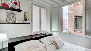 A bedroom with a bed, a bedside table with a lamp, a decorative shelf with a vase and flowers, and a view of the outside through a window. - Marco Polo Apartment by Wonderful Italy (Venecia)