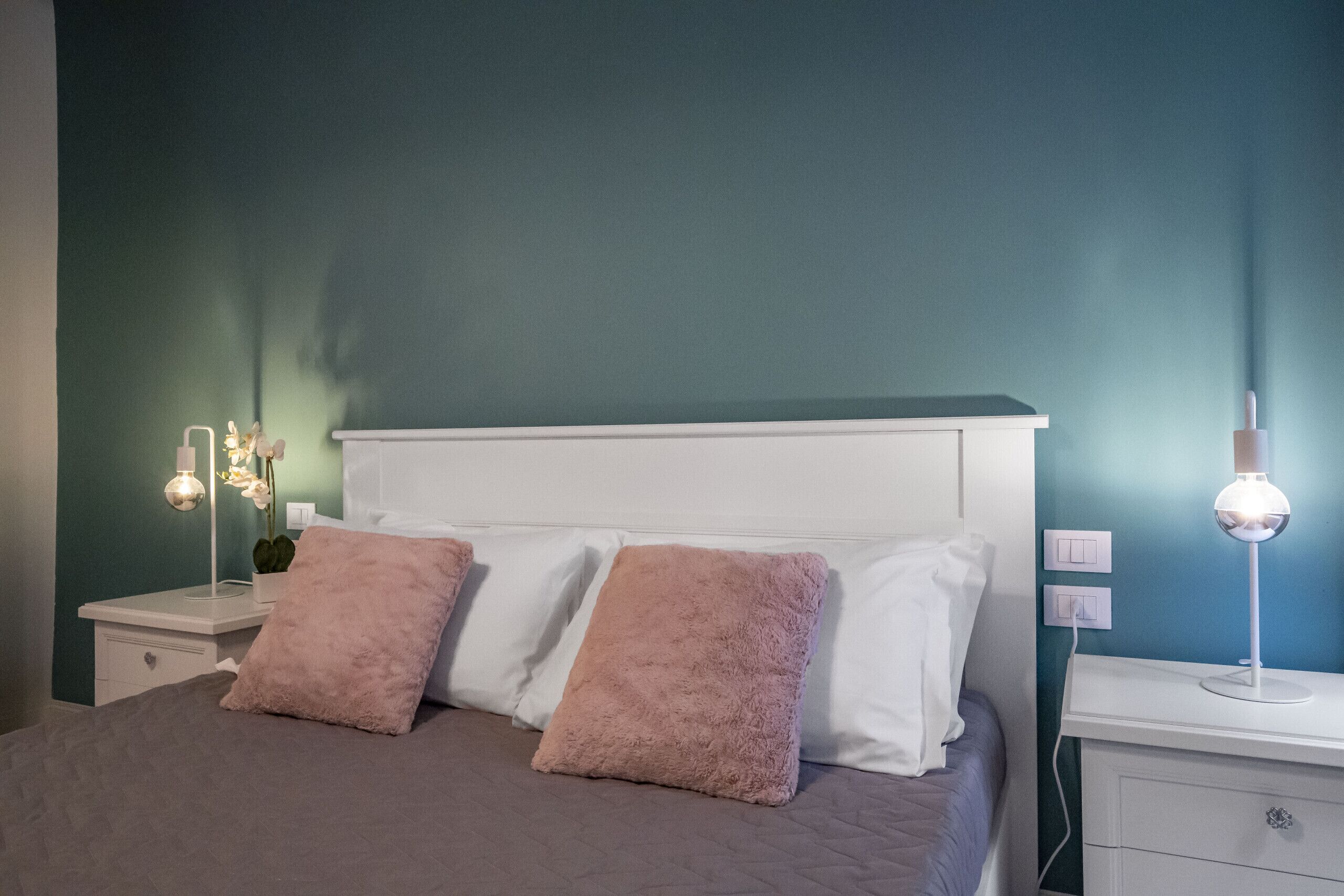 A neatly made bed with two pillows, a bedside table with a lamp, and a wall switch.