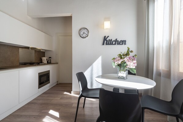 A modern kitchen with a clock, a 'Kitchen' sign, a dining table with chairs, and a vase with flowers.