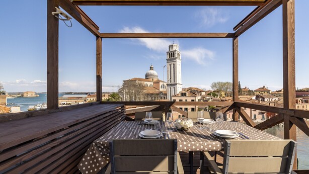 A wooden deck with a table set for two, overlooking a body of water and a historic building with a bell tower. - Skyline Penthouse by Wonderful Italy (Venecia)