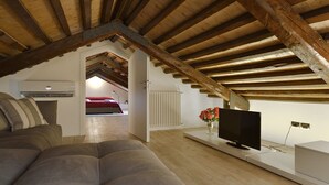 A loft bedroom with a sloped ceiling, wooden beams, a bed, a TV on a white stand, and a radiator. - Skyline Penthouse by Wonderful Italy (Venecia)