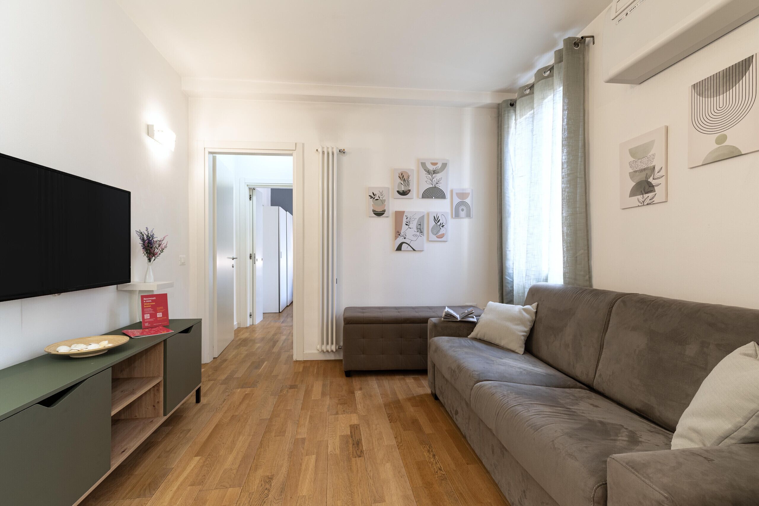 A modern living room with a flat-screen TV, a grey sofa, wooden flooring, and a hallway leading to a door.