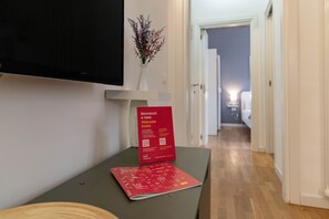 Un couloir avec un téléviseur à écran plat fixé au mur, un petit vase avec des fleurs sur une table et une pancarte rouge indiquant « Bienvenue ».