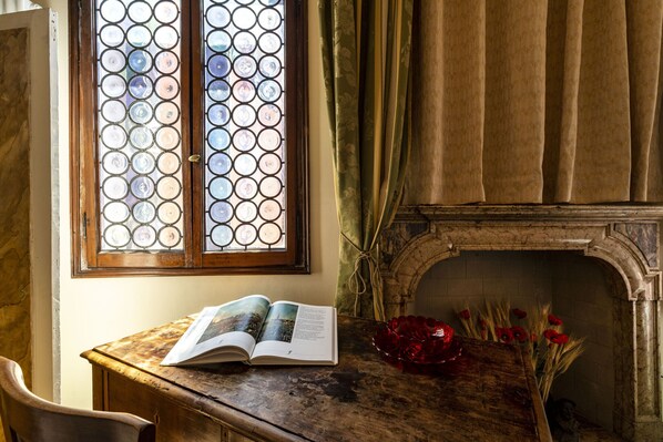 Una habitación con un escritorio de madera, un libro abierto, un jarrón con flores rojas, una chimenea y una ventana con un diseño decorativo en el vidrio.