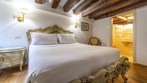 Un dormitorio con una cama grande, vigas de madera en el techo, dos apliques, una mesita de noche y una puerta que da al baño.