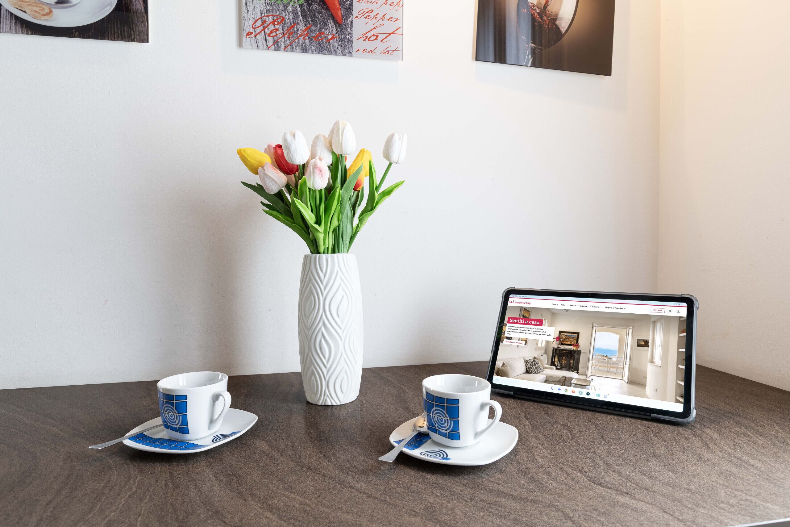 A tablet displaying a room layout, two cups with saucers, and a vase of tulips on a table.