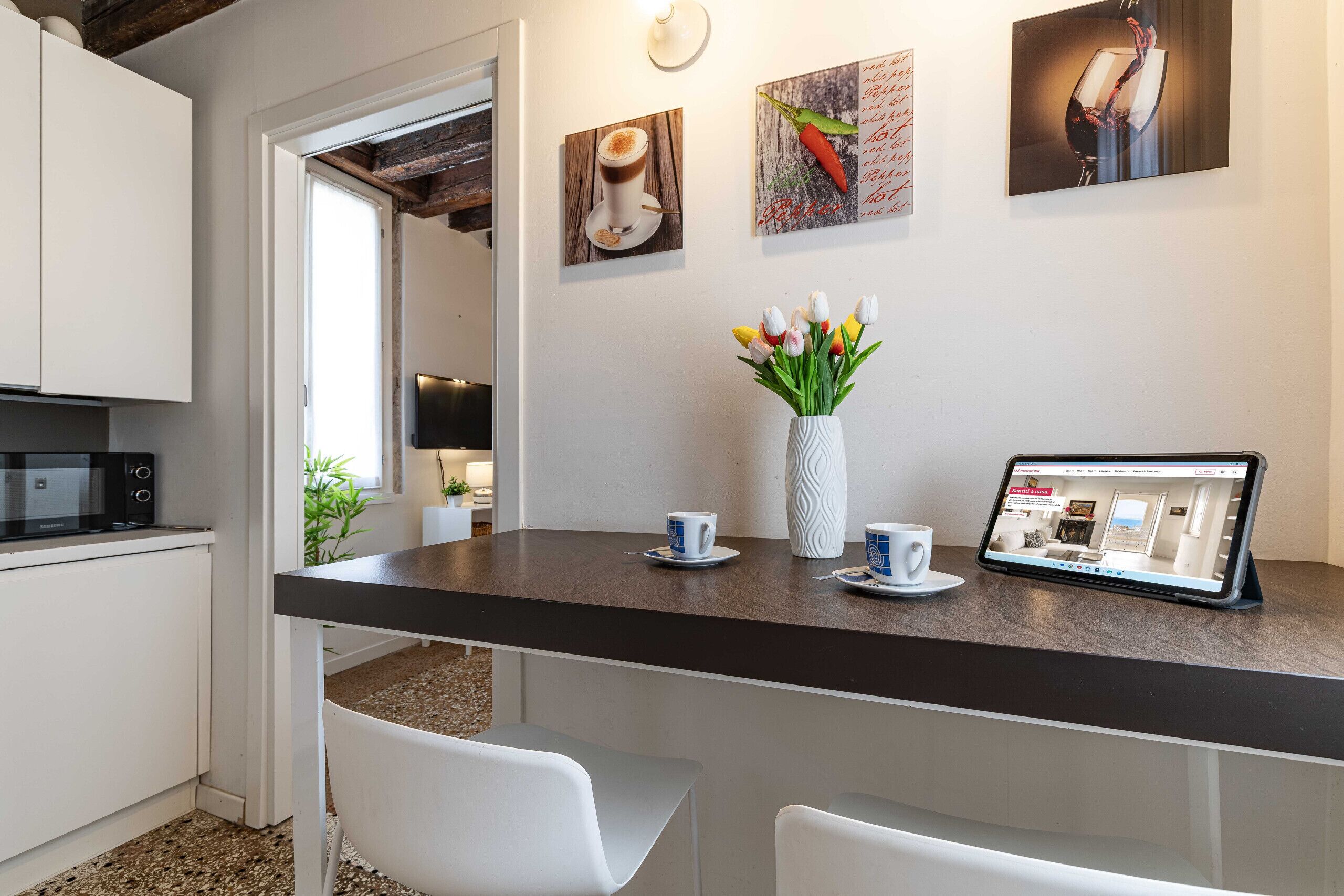 Una cocina moderna con encimera oscura, sillas blancas, un florero blanco con tulipanes y una tableta mostrando un sitio web.