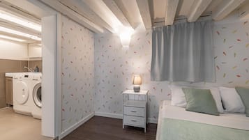 A compact bedroom with a washing machine, a bedside table with a lamp, and a bed with pillows.
