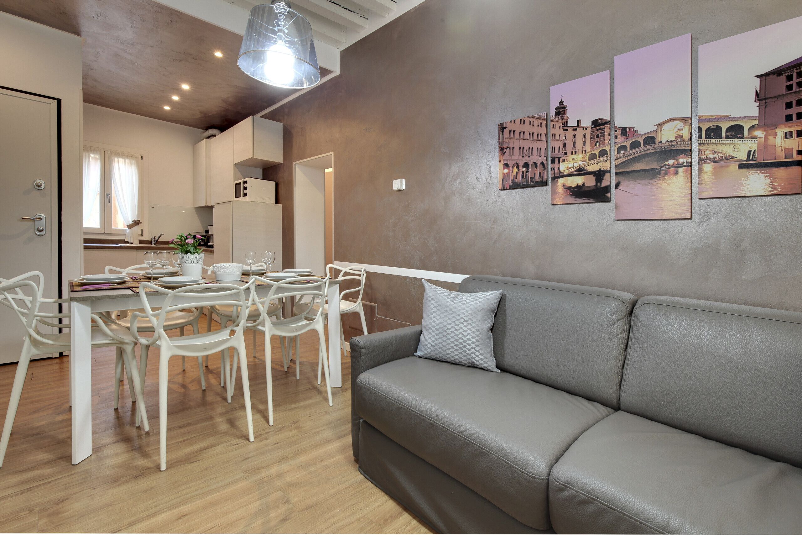 A modern living room with a grey leather sofa, a dining table with white chairs, and a kitchen in the background.
