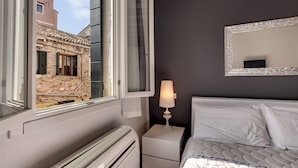 A bedroom with a bed, a nightstand with a lamp, a window with a view of a building, and a radiator.