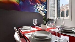 A dining table set with white plates, glasses, and cutlery, a vase with white flowers, and a colorful abstract painting on the wall.