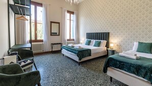 Une chambre d’hôtel avec un lit, une chaise, une table, un radiateur, une fenêtre avec des rideaux et un lustre.
