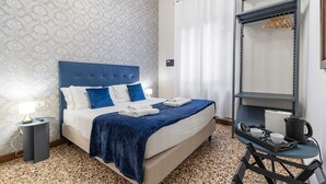 Habitación de hotel con una cama, una mesita de noche, un carrito con tazas y un hervidor, y un armario con ropa colgada.
