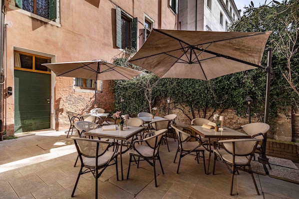 An outdoor patio with tables and chairs, umbrellas, and a building in the background. - Centopietre by Wonderful Italy - Camera Blue Moon (Venecia)