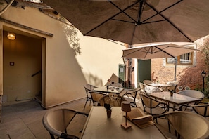 Un patio al aire libre con mesas y sillas, un parasol grande y una puerta que da a un espacio interior.