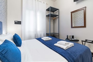 A hotel room with a bed, a blue bedspread, white pillows, a blue pillow, a towel, a mirror, a small table with a tray, and a framed picture on the wall. - Centopietre by Wonderful Italy - Camera Blue Moon (Venecia)