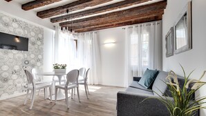 A modern living room with a grey sofa, white chairs, a dining table, and a wooden ceiling.