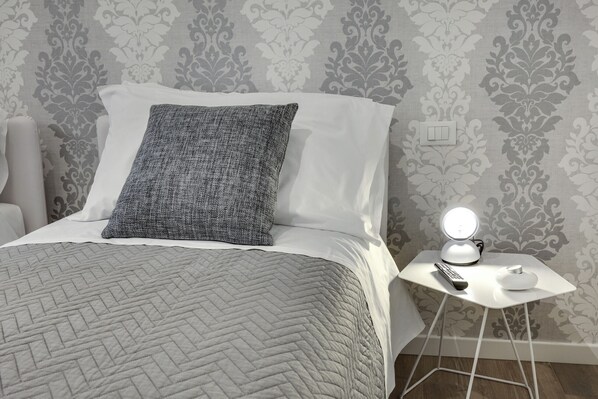 Een net opgemaakt bed met een gewatteerde deken, een grijs kussen met structuur en een wit kussen. Daarbij staat een klein wit nachtkastje met een lamp en een afstandsbediening.