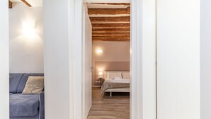 En smal gang der fører til et soveværelse med en seng og et natbord, en blå sofa med pude, og et træloft.