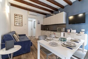 Een moderne woonkamer met een blauwe bank, een eettafel gedekt met borden en glazen, een aan de muur bevestigde televisie, en een keuken met kasten en apparatuur.