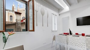 A dining area with a table set for four, a large window with a view of a building, and a wall-mounted television. - Bellini Design by Wonderful Italy (Venecia)